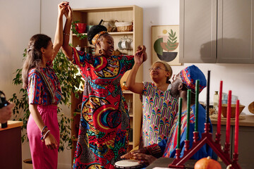 Happy mother and two daughters in national costumes holding by hands and dancing in front of man beating drum during kwanzaa celebration