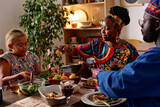 Fototapeta Na drzwi - Young African American woman passing baked corn to her daughter by festive table served with homemade food for kwanzaa dinner