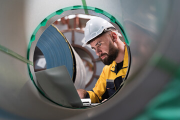 Male factory worker using laptop computer inspecting quality rolls of galvanized or metal sheet in in aluminum material warehouse. Male worker working in warehouse during distribution process