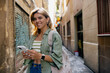 Cute blond woman with wonderful smile wearing green shirt and holding backpack is using smartphone on background od old city streets 