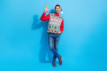 Sticker - Full length photo of good mood positive guy wear christmas hat print waistcoat waving palm say hi isolated on blue color background