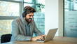 © Bojan - Young middle eastern businessman working on laptop computer wearing headphone at modern office. Smiling business man wearing headset for video conference, communication, education and e-learning.