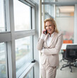 © Bojan - Smiling businesswoman using her phone in the office. Small business entrepreneur looking through window and smiling while communicating with her office colleagues.