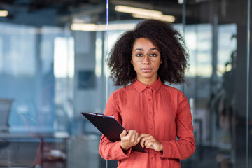 Portrait of serious confident business woman with documents inside office, successful female worker looking thoughtfully and concentrated at camera, african american woman with curly hair