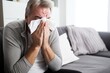 Gray-haired, elderly man sneezes into a handkerchief while sitting on the sofa in the living room. Concept of self-isolation at home for cold and flu symptoms