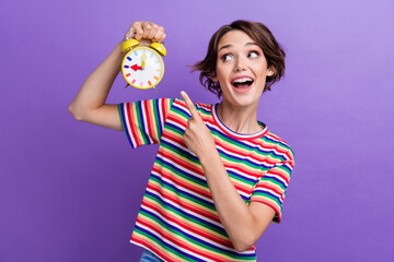 Poster - Photo of glad cute girl dressed stylish striped clothes looking alarm clock isolated on purple color background