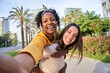© CarlosBarquero - Multiracial lesbian young couple taking selfie portrait outdoor sunny day. Beautiful Caucasian woman giving piggyback ride to nice African girl on city street. Smiling female posing for photo happy.