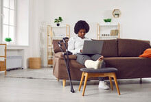 African American Woman With Fractured Leg Sitting On Sofa And Using Laptop Computer. Beautiful Girl With Plastered Leg Sitting On Couch Working Online While Recovering At Home