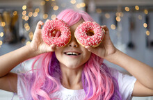 Close Up Portrait Of A Little Smiling Girl With Pink Hair And Two Appetizing Donuts In Her Hands, On A Kitchen Background. Vanilla Girl. Kawaii Vibes.