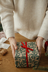 Wall Mural - Woman holding stylish christmas gifts with red ribbon at wooden table with festive decorations in scandinavian room. Merry Christmas and happy holidays!