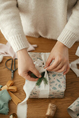 Woman wrapping stylish christmas gift with ribbon and fir branch on wooden table with festive decorations in modern decorated scandinavian room. Merry Christmas! Hands packing present close up