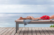 Les jambes d'un  homme en maillot de bain sur transat en terrasse, serviette pend du transat, mer et ciel  en arrière-plan. Instant de détente au bord de l'eau