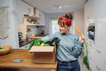 Smiling Woman Eating Fresh Carrot From Her Healthy Food Delivery Box. Box With Vegetables And Phone With Veganuary App On The Kitchen Table. Start Of A Healthy Life Concept. Online Home Food Delivery.