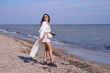 Wall Mural - Portrait of attractive brunette girl with long hair posing on a deserted beach. She wears white shorts, a top, a long white cape. She is looking to the camera.