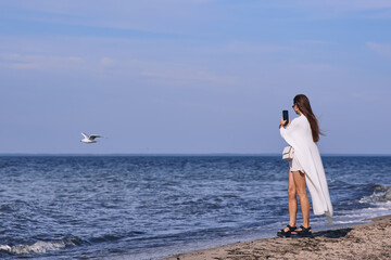 Wall Mural - Portrait of attractive brunette girl with long hair posing on a deserted beach. She wears white shorts, a top, a long white cape. She is looking to the camera.