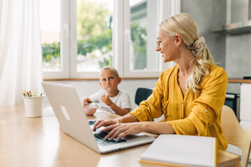 Caucasian woman is working from home on laptop and sitting in the kitchen with her son.