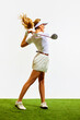 Beautiful, sportive, blonde, young girl with slim, fit body playing golf on grass, teeing off isolated over white background. Concept of sport, hobby, beauty and fashion, relaxation, game