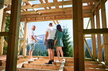 Man designer with plan showing to couple the process of constructing wooden framed house. Back view of investors inspecting their future dwelling. Concept of contemporary ecological construction.