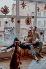 Beautiful young couple in love at the window hugging, next to their dog