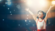 Little Asian gymnast joyfully raising arms under spotlight in a gym.