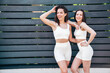 Two young beautiful smiling female in trendy summer white cycling shorts and top clothes. Sexy carefree women posing in street at sunny day. Positive models having fun. Cheerful and happy. Near fence