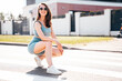 Young beautiful smiling female in trendy summer blue cycling shorts and tank top clothes. Carefree woman posing in street. Positive model having fun. Cheerful and happy. In sunglasses. Sits on asphalt
