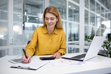 Successful mature female financial accountant at workplace inside office, fills data in table, works with documents and papers, uses laptop and calculator for paperwork.
