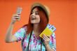 Close up of woman in straw hat with flight tickets and passport, taking selfie on smartphone smiling, isolated on orange background. Portrait of female traveler.