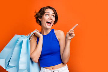 Wall Mural - Portrait of attractive impressed girl hold store bags look direct finger empty space advert isolated on orange color background