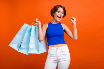 Wall Mural - Portrait of delighted nice lady hold store mall bags raise fist success isolated on vivid orange color background