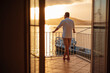 Handsome middle aged barefoot man in white shirt and shorts standing on balcony and looking at sea. Beautiful sunset on background