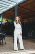 © DSMT - A young stylish woman in a white classic suit with a smile on her face is standing on the terrace of a summer cafe.