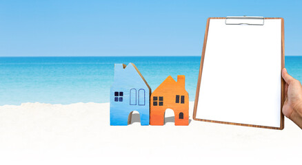 Wall Mural - Wooden miniature house with blank clipboard in girl hand on tropical beach, outdoor day light, real estate and property business concept