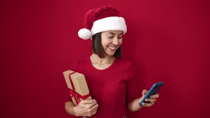 Canvas Print - Young beautiful hispanic woman wearing christmas hat using smartphone holding gift over isolated red background