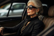 Business elderly lady in black stylish clothes and with jewelry driving a car, confident rich senior Caucasian woman driver in sunglasses