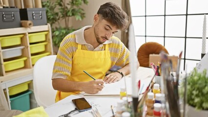 Poster - Stressed young arabian man artist, looking at smartphone while drawing in notebook, grapples with art stress in studio.