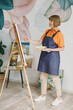© ViDi Studio - Full body elderly fun happy artist woman 50 years old wearing casual clothes stand near easel with painting artwork paint look aside spend free spare time in living room indoor. Leisure hobby concept.