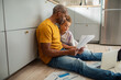 © Marko Geber - Father and daughter review documents together while seated on the kitchen floor with a laptop nearby