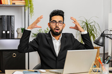 Excited amazed Indian businessman freelancer looking shocked surprised wow showing hands brain head explosion gesture working at modern home office workplace desk. Worried male Arabian in formal suit