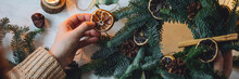 Young Woman In Knitted Sweater Doing Handmade Christmas Wreath From Natural Materials: Fir Tree Branches, Dry Citrus Slices, Cones, Cinnamon Sticks. Wooden Table, Cozy Light, Home Atmosphere. Banner