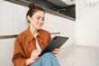 Portrait of pretty young woman at home, sitting on kitchen floor, looking at digital tablet, using gadget, reading on device