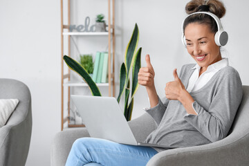 Sticker - Mature woman with laptop and headphones showing thumb-up at home