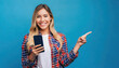 Young happy latin woman holding mobile phone pointing aside isolated on blue background. Smiling female model holding cellphone using cell presenting advertising new trendy shopping sale in app
