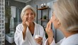 Attractive mid age older adult 50 years old blonde woman wears bathrobe in bathroom applying nourishing antiage face skin care cream treatment, looking at mirror doing daily morning beauty routine