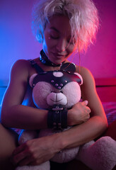 Wall Mural - sexy girl in leather in a bdsm accessory on the bed with a cute teddy bear with emotions and in neon light