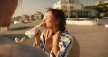 Close-up Shot Of A Brown-haired Girl In A White Blue Shirt Eating A Hot Dog In The Summer On A Modern Beach In The Morning