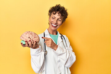 Wall Mural - Doctor holding a brain model on yellow studio smiling and pointing aside, showing something at blank space.