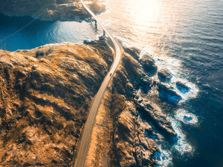 Wall Mural - Aerial view of bridge, car, sea with waves and mountains at sunset in Lofoten Islands, Norway. Landscape with beautiful road, water, rocks and stones, golden sunlight. Top view from drone of highway