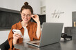 Portrait of young self-employed woman, replying to customers on smartphone, using laptop to check orders, sitting at home