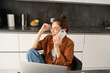 Image of beautiful modern woman working from home, studying in kitchen with laptop, talking on smartphone, calling someone on telephone